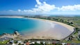 A coastal town with a sandy beach, clear blue waters, and rolling hills in the background.