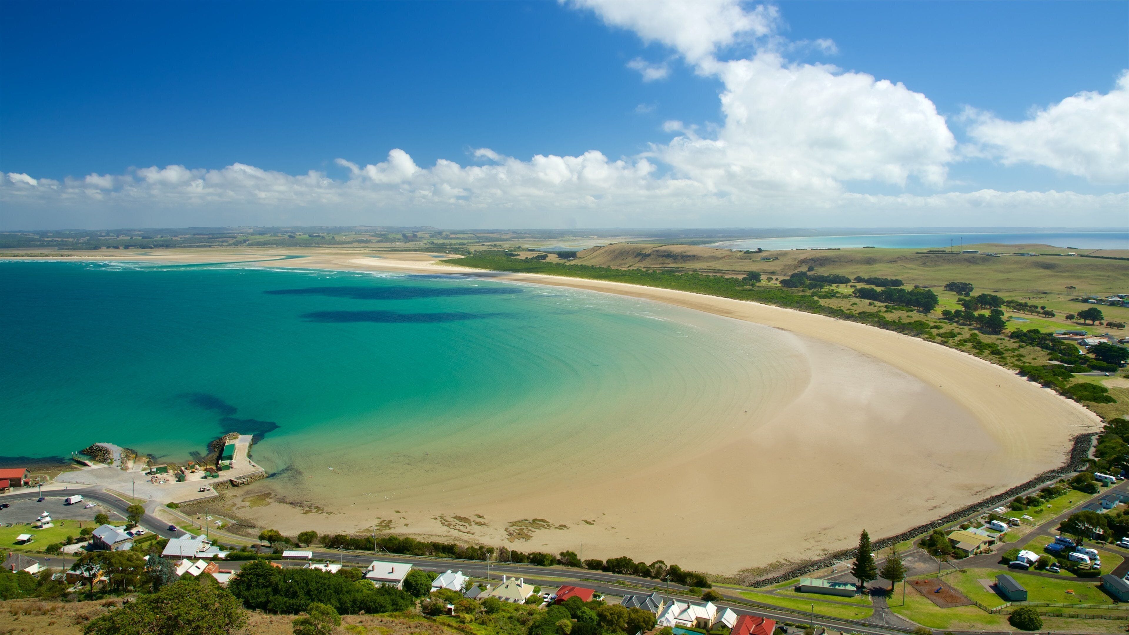 A coastal landscape with a sandy beach, clear turquoise waters, and a small town with houses and roads.