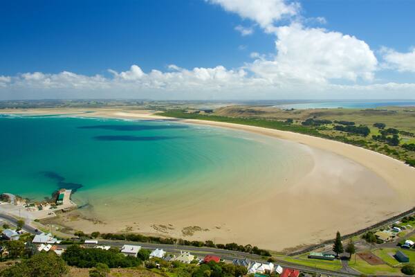 A coastal landscape with a sandy beach, clear turquoise waters, and a small town with houses and roads.