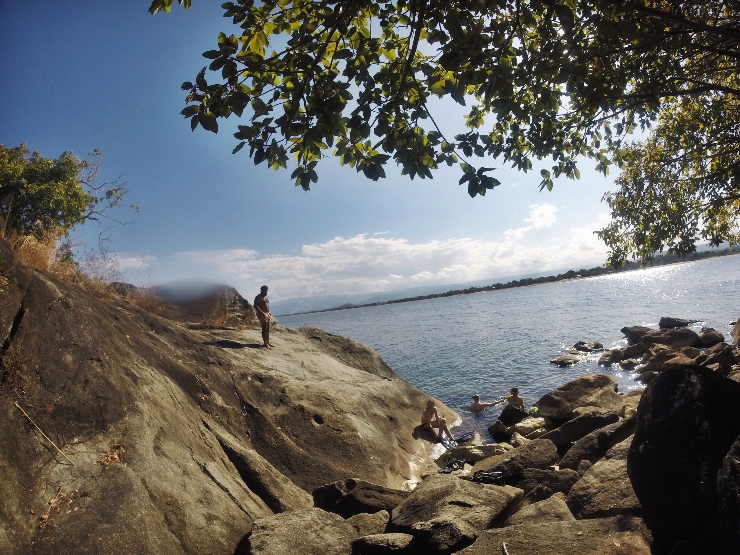 A rocky shoreline with people enjoying the water, a clear sky, and distant mountains.