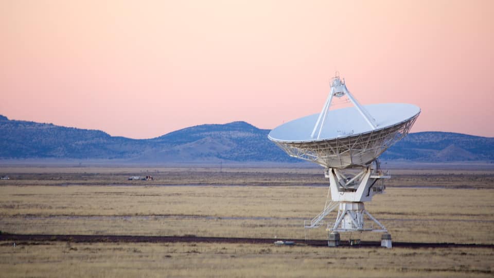 A large satellite dish in a flat, open field with mountains in the background.