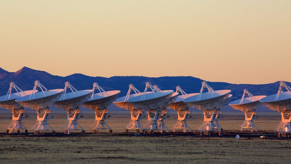 A series of large radio telescopes in a flat, open field with mountains in the background.