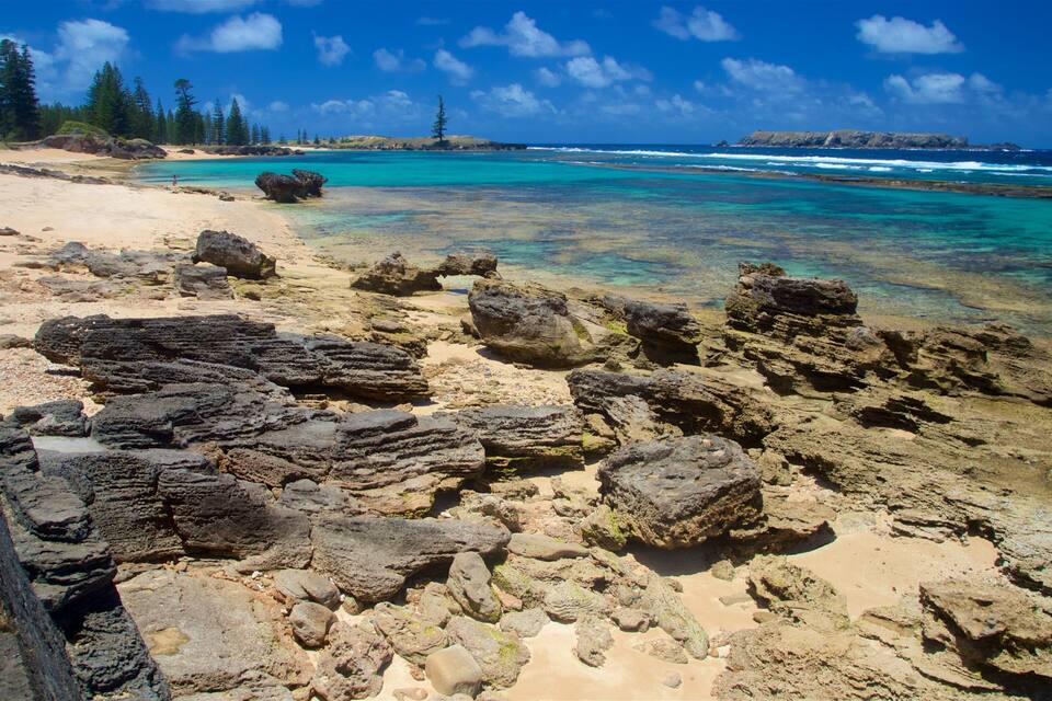 A rocky shoreline with clear turquoise water, sandy beach, and distant mountains under a blue sky.
