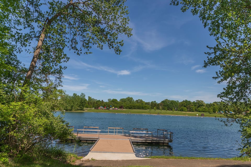 A lake with a wooden dock extending into the water, surrounded by trees and a clear blue sky.