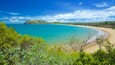 Paisagem costeira com mar turquesa, praia de areia e colinas verdes.