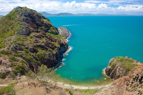 Un paysage côtier caractérisé par une falaise rocheuse, des eaux turquoise et limpides, et des montagnes à l’horizon.