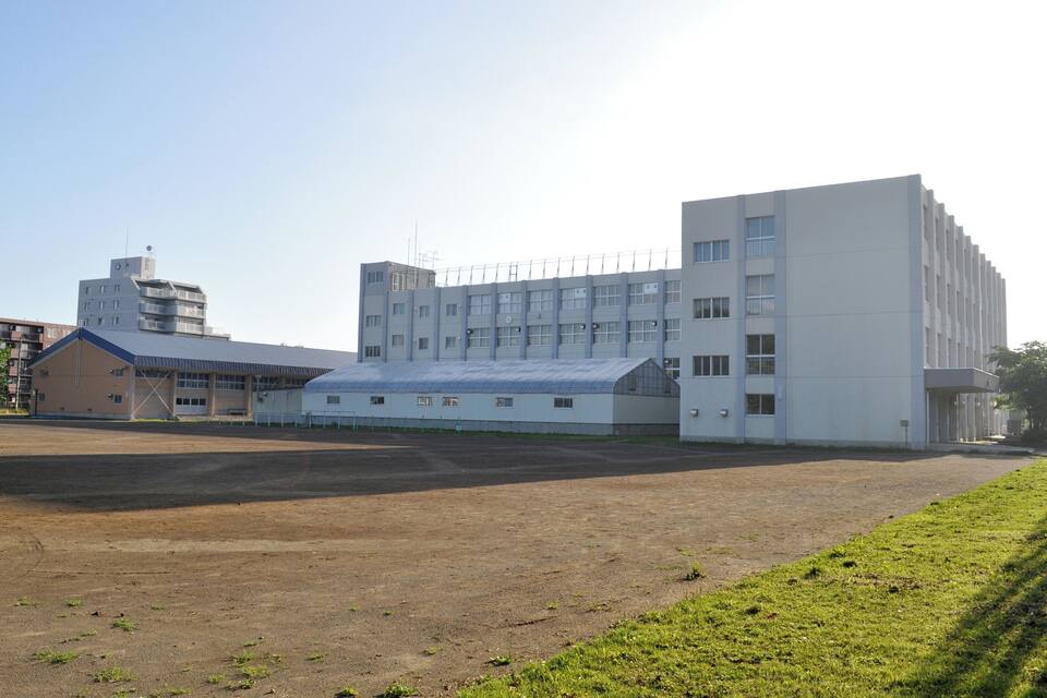 A large industrial building with a flat roof and multiple windows, surrounded by a grassy field and a dirt area.