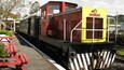 Un train d’époque, avec une partie avant jaune et une partie arrière rouge, est garé dans une gare où se trouvent des bancs rouges et de la verdure.