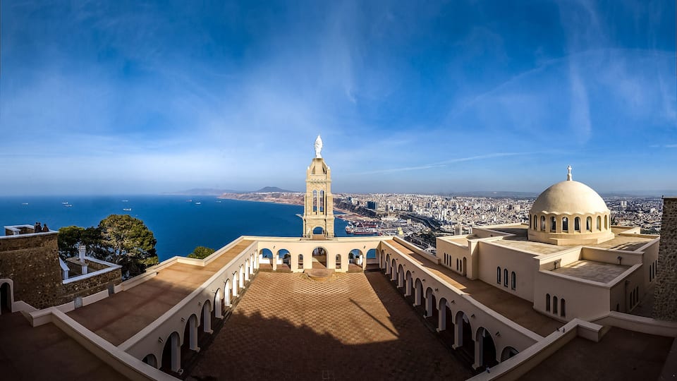 A coastal cityscape with a prominent tower and dome, a clear blue sky, and the sea in the background.
