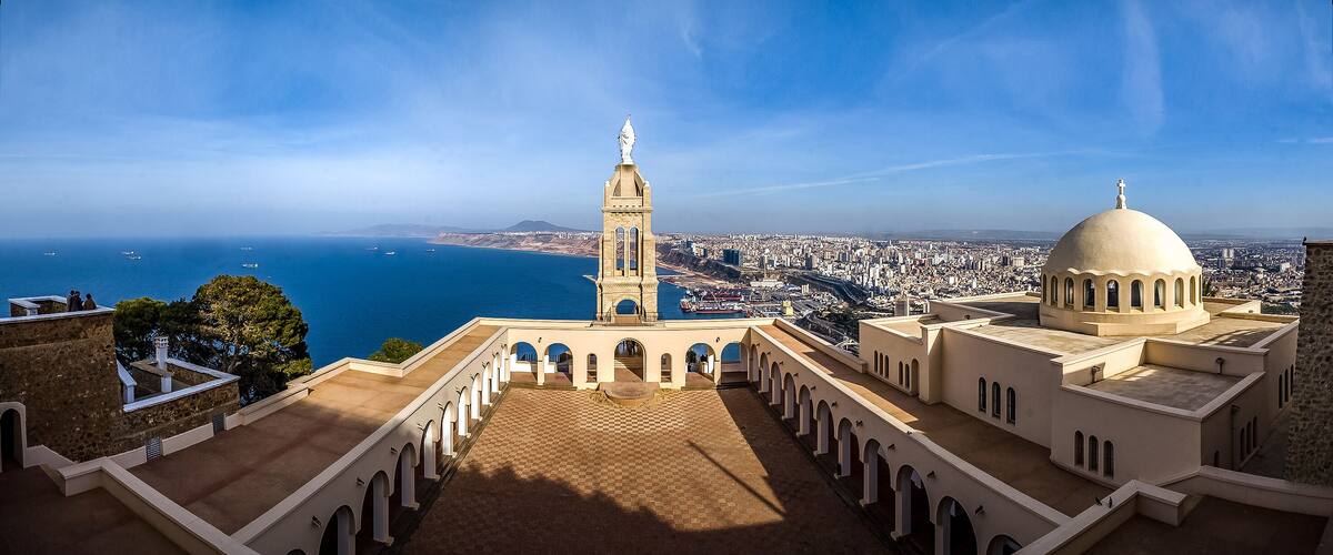 A coastal cityscape with a prominent tower and dome, a clear blue sky, and the sea in the background.
