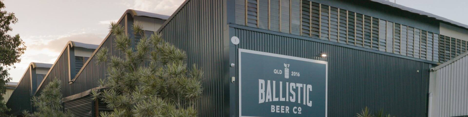 A building with a sign that reads "Ballistic Beer Co." There are bicycles parked in front, and people are sitting at tables outside.