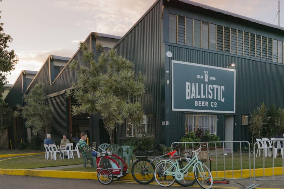 A building with a sign that reads "Ballistic Beer Co." There are bicycles parked in front, and people are sitting at tables outside.