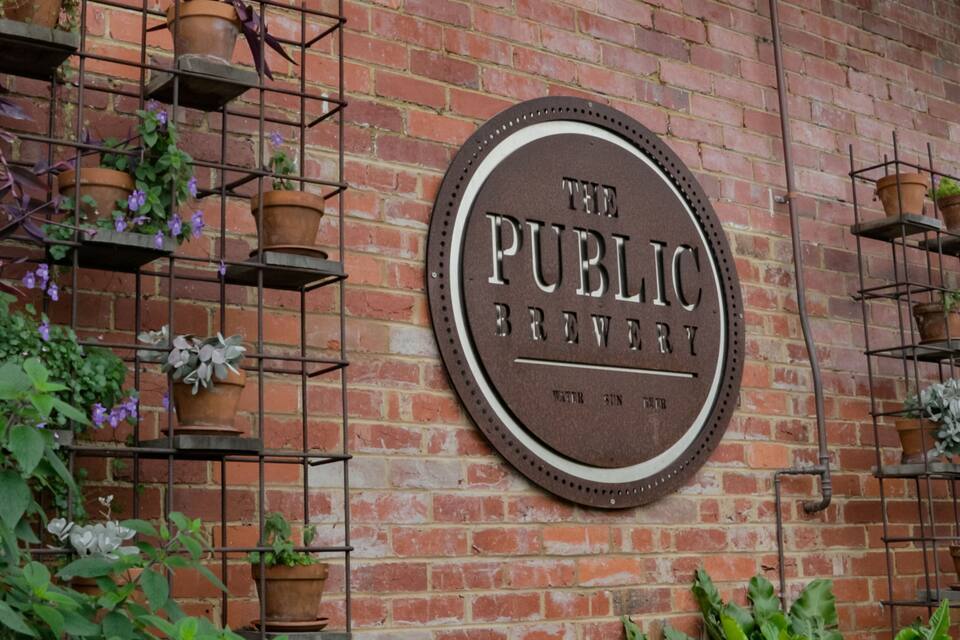 A brick wall with a circular sign that reads "The Public Brewery" and a metal plant holder with various plants.