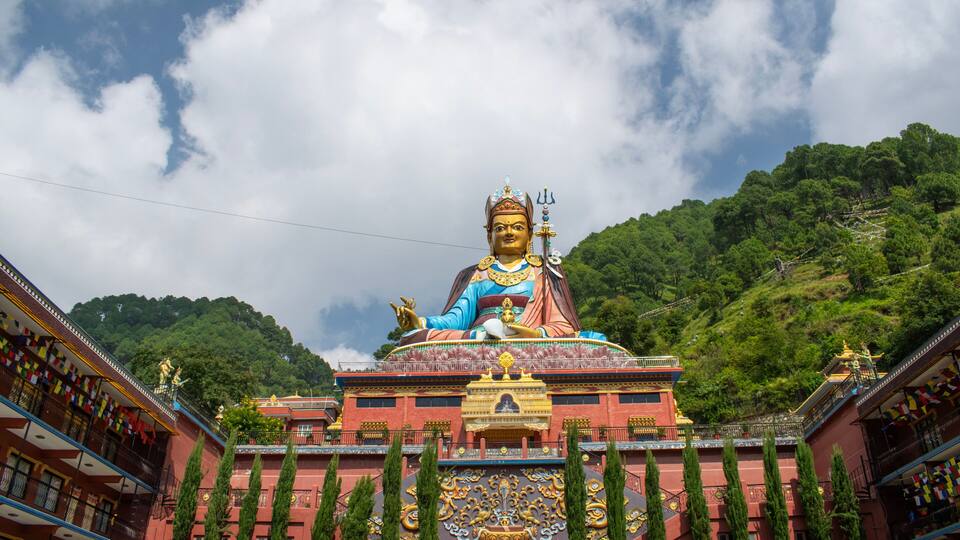A large statue of a deity with a golden halo, seated on a red and gold pedestal, surrounded by traditional buildings and lush green hills.