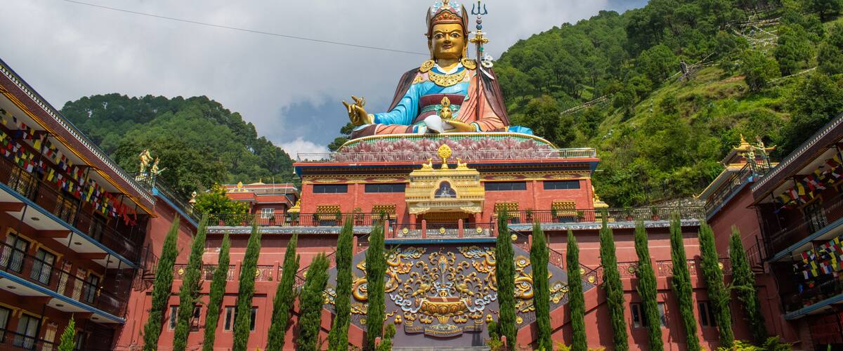 A large statue of a deity with a golden halo, seated on a red and gold pedestal, surrounded by traditional buildings and lush green hills.