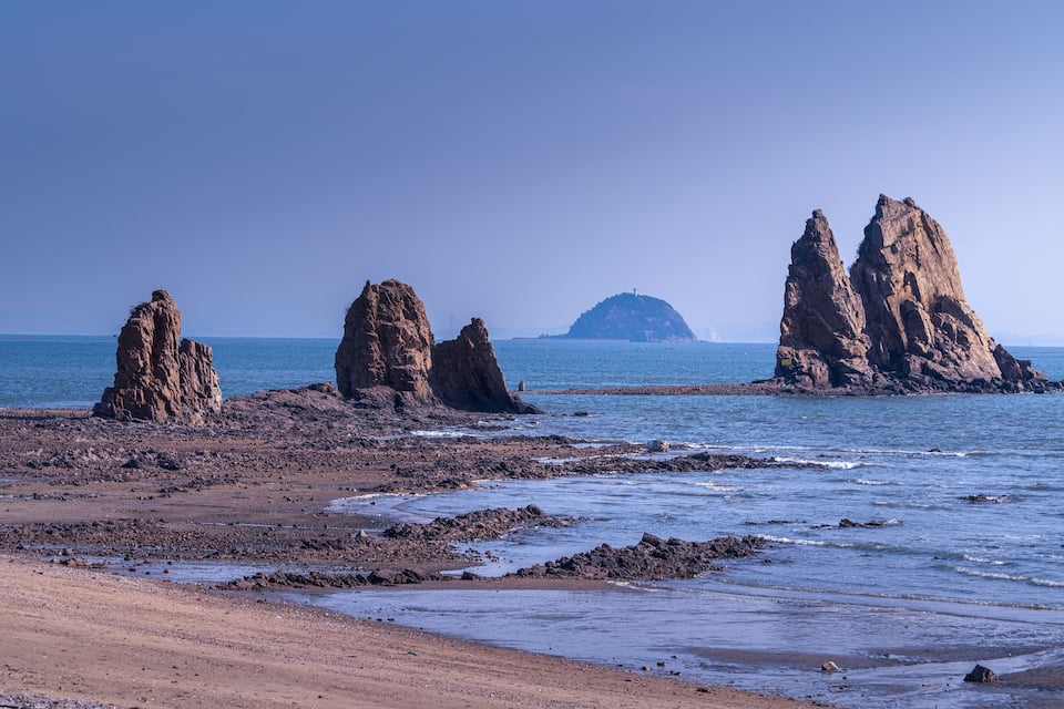 Litoral rochoso com formações rochosas imponentes e uma ilha ao longe.