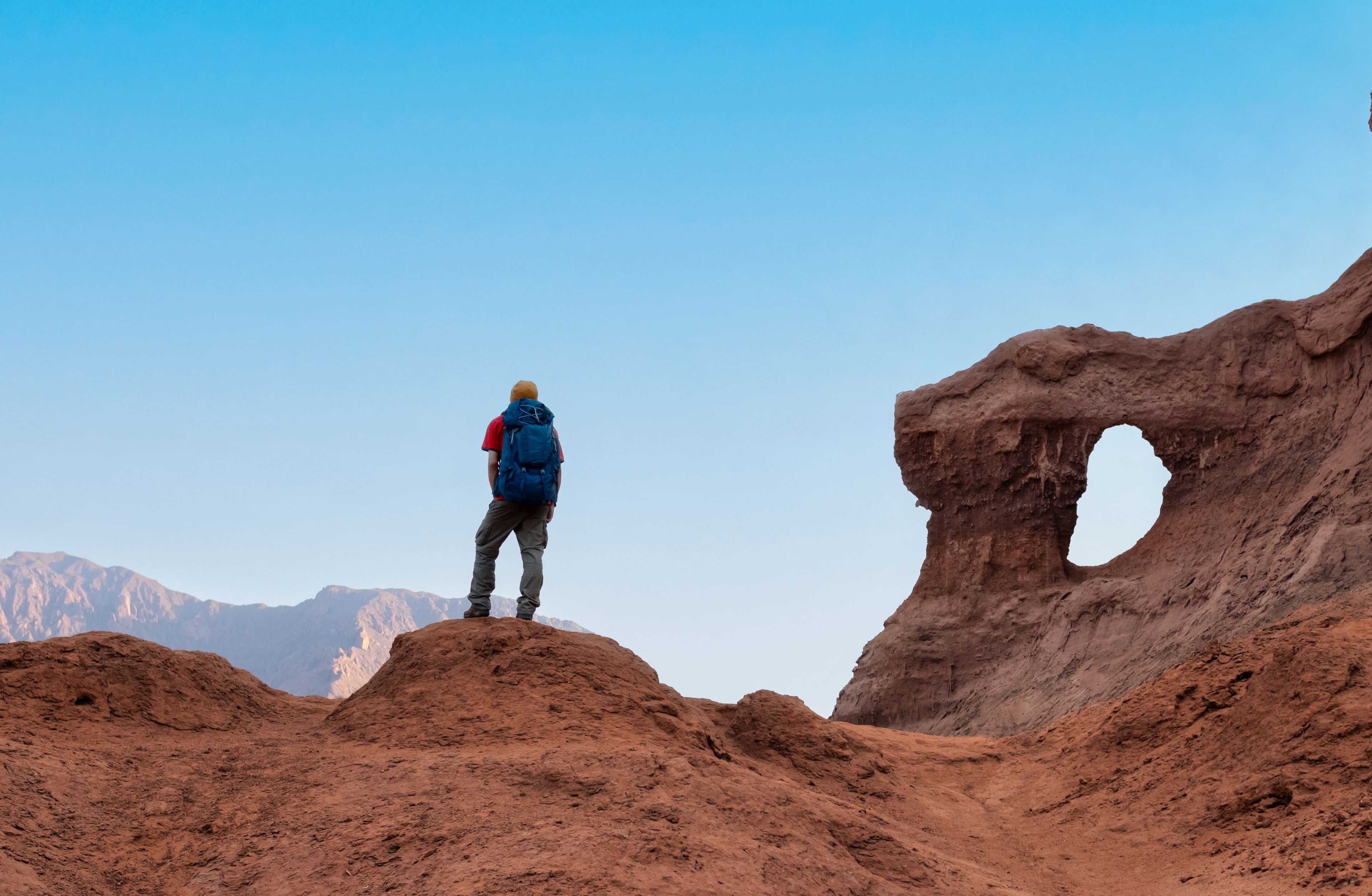 A person standing on a reddish-brown rocky terrain with a large rock formation featuring a natural arch.