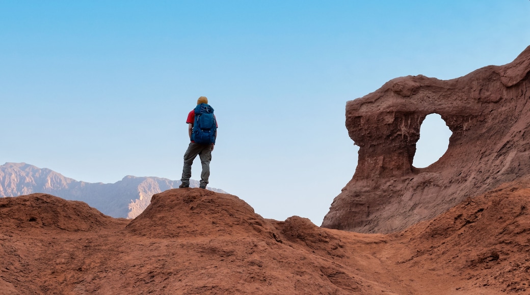 A person standing on a reddish-brown rocky terrain with a large rock formation featuring a natural arch.