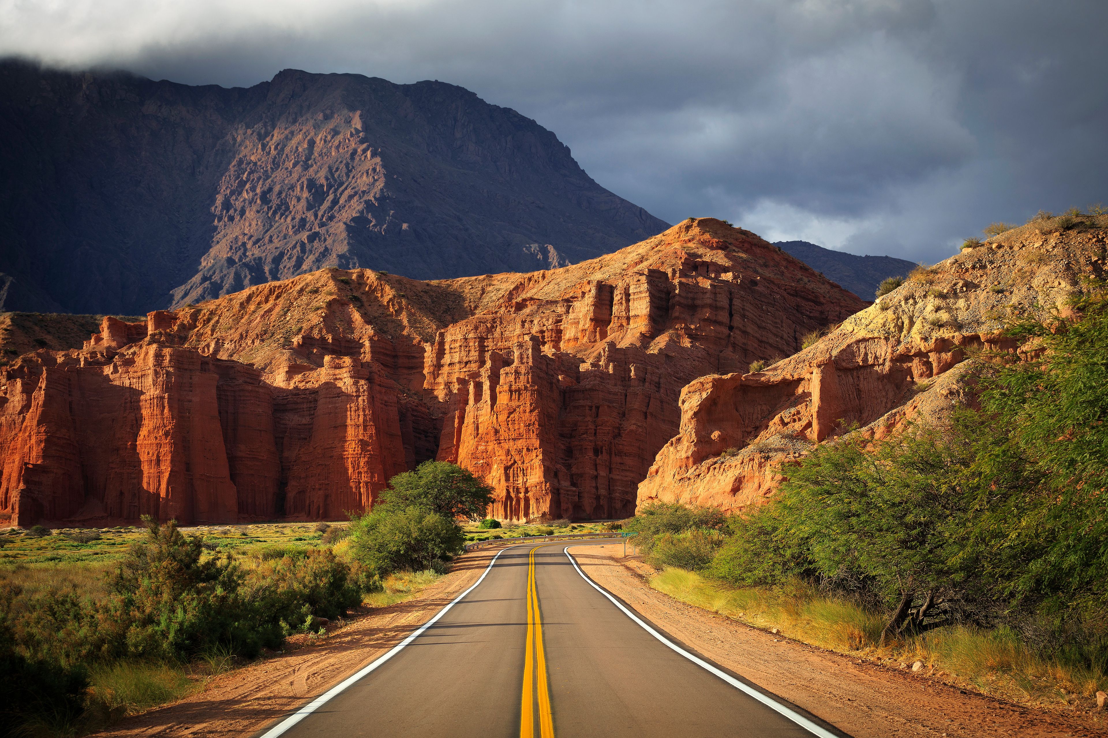 A road leading through a canyon with red rock formations and a mountain in the background.