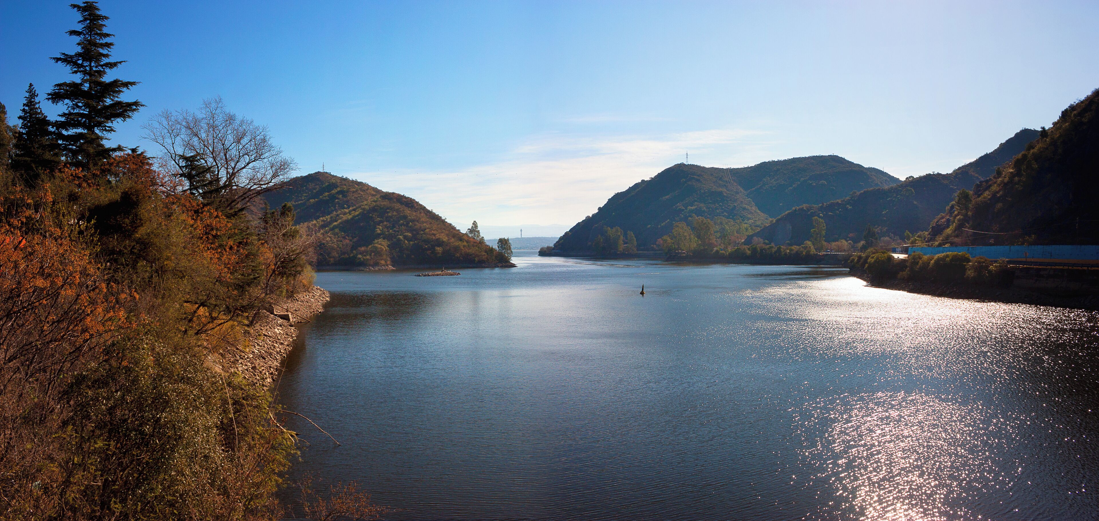 A calm river flanked by hills with autumn foliage, a clear sky, and a distant bridge.