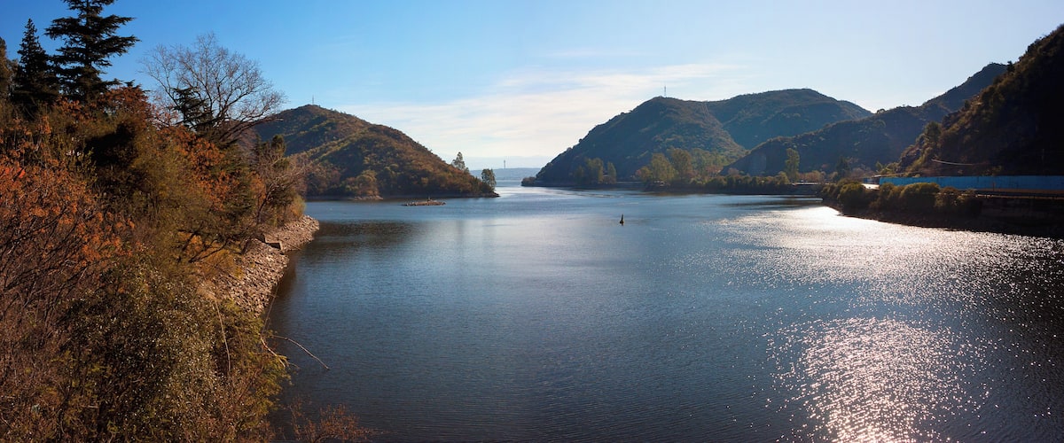 A calm river flanked by hills with autumn foliage, a clear sky, and a distant bridge.