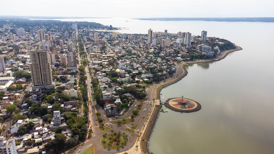 An aerial view of a coastal city with dense urban development, a large body of water, and a distinctive circular landmark near the shore.
