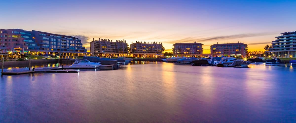A marina at dusk with boats docked, waterfront buildings, and a sunset sky.