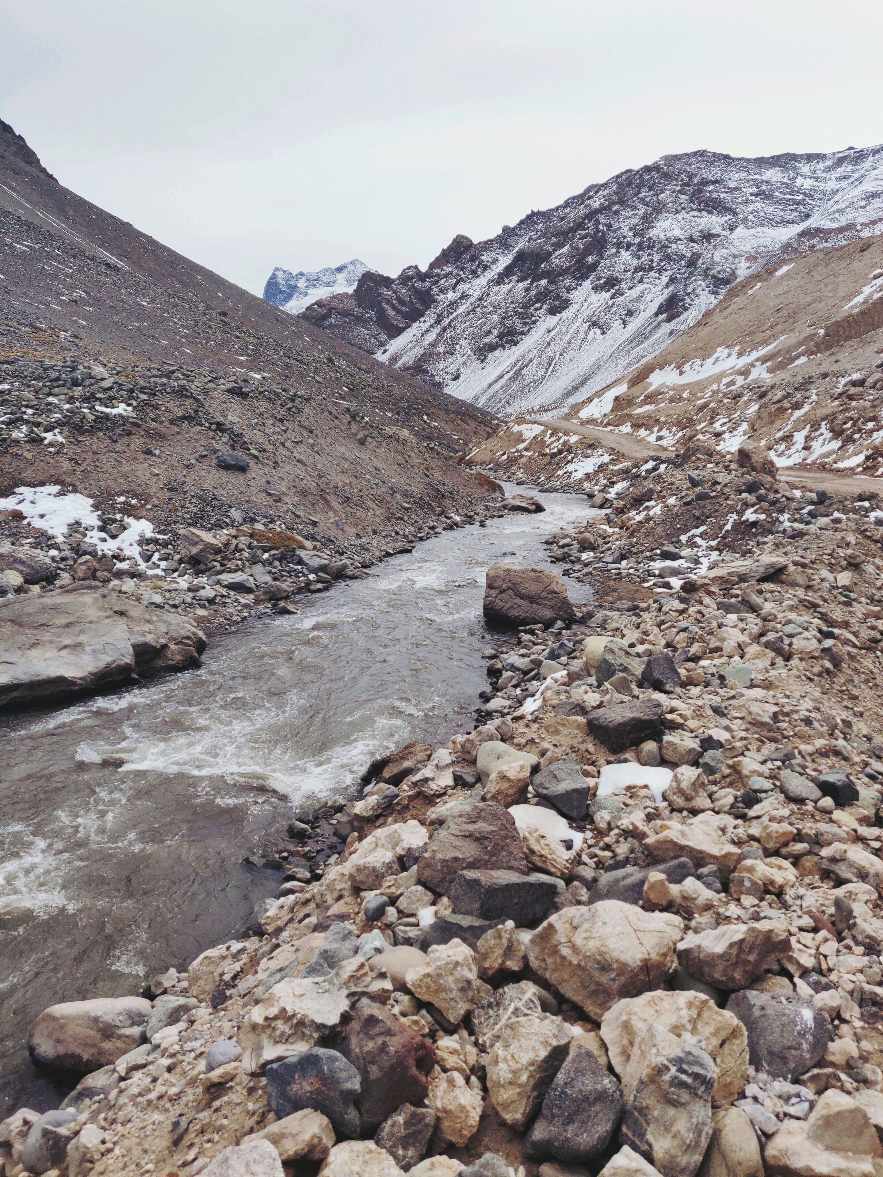 A rocky riverbed with snow patches and mountains in the background.