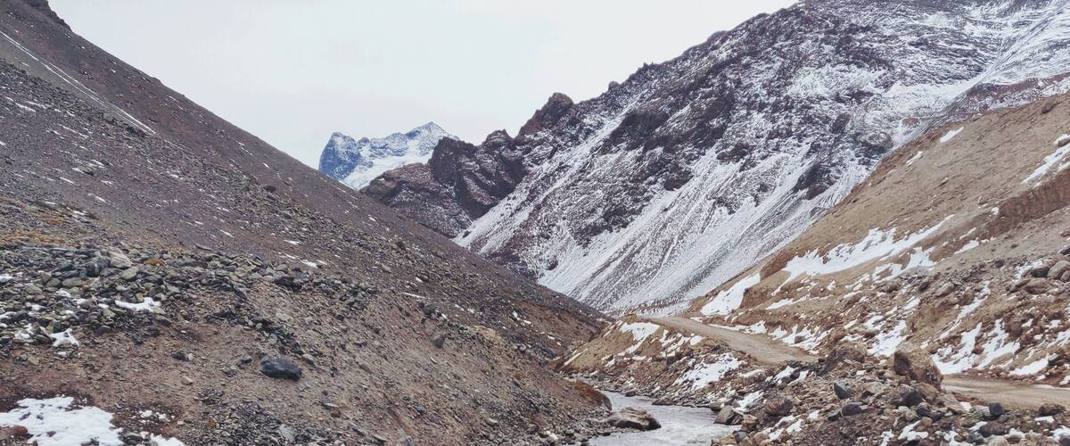 A rocky riverbed with snow patches and mountains in the background.
