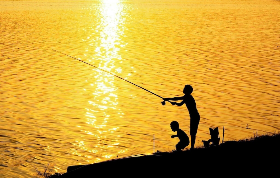 A silhouette of a person fishing with a child beside them, against a sunset over a body of water.