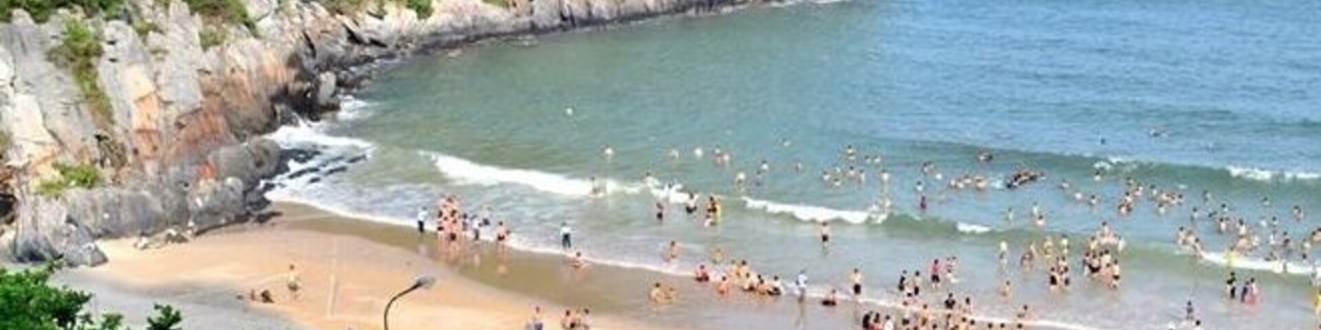 A crowded beach with people enjoying the water, surrounded by greenery and rocky cliffs.