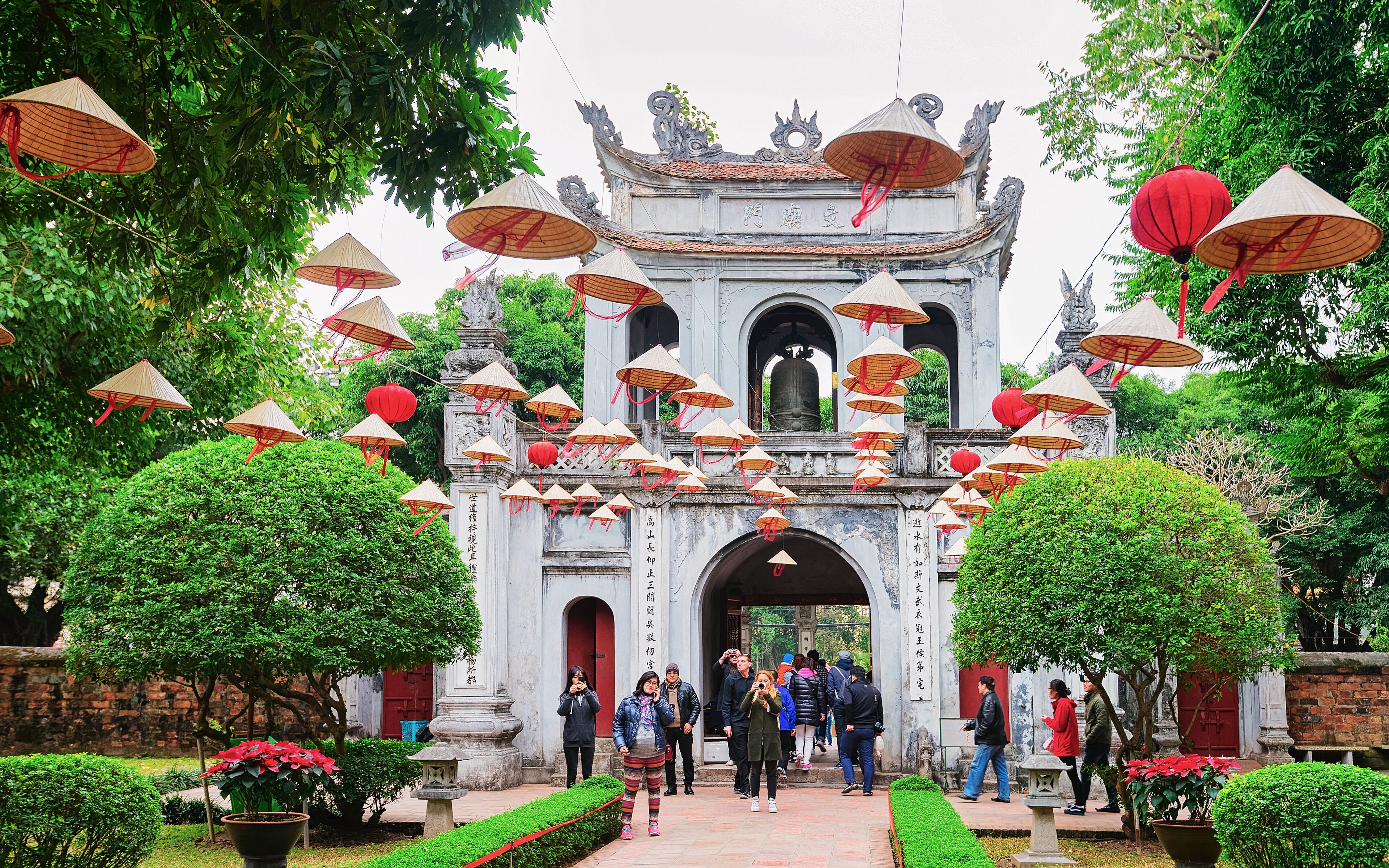A traditional gate with hanging conical umbrellas and red lanterns, surrounded by lush greenery and visitors.