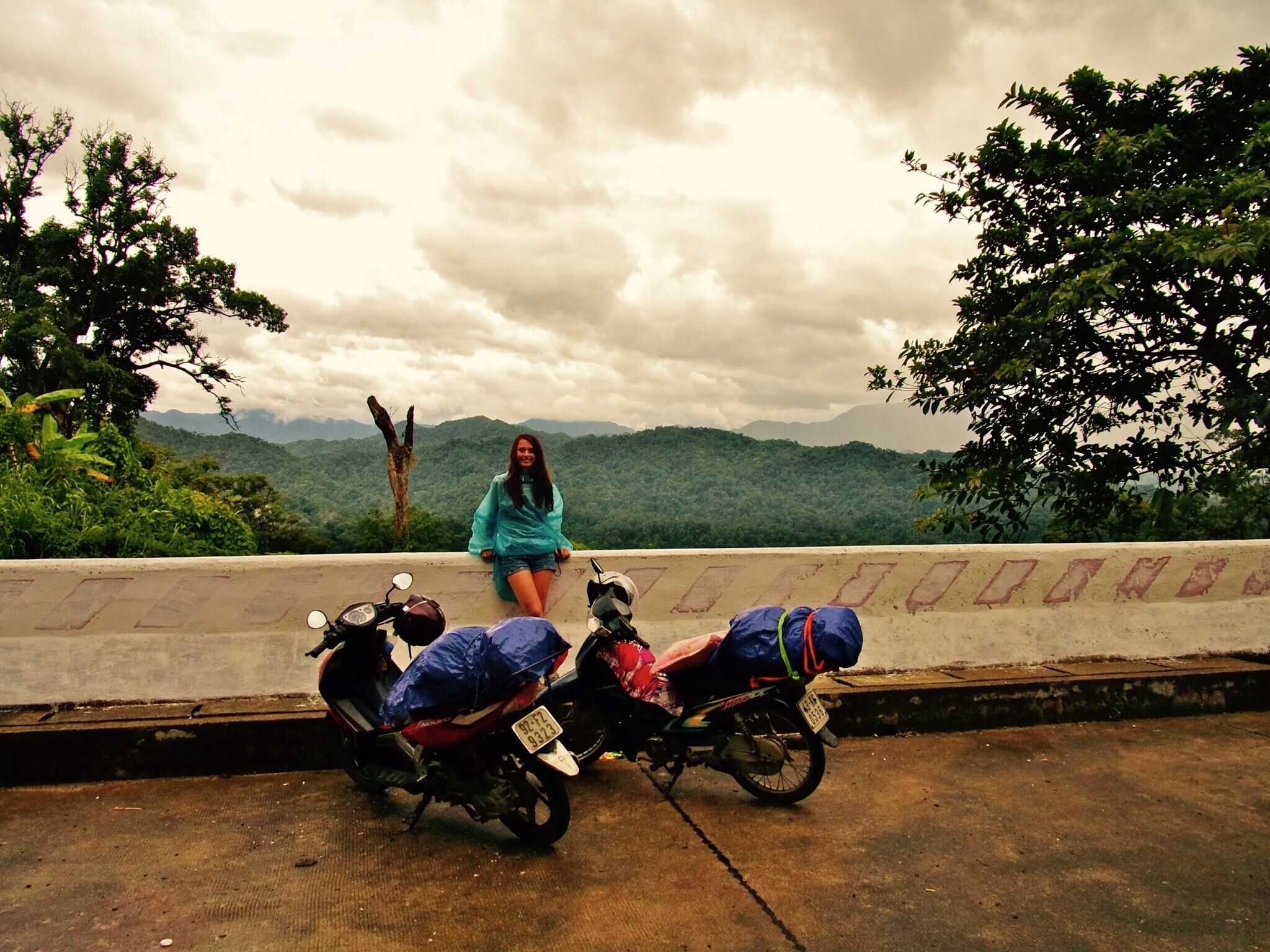 Una mujer sentada en un saliente con vista a un paisaje montañoso y dos scooters estacionados.