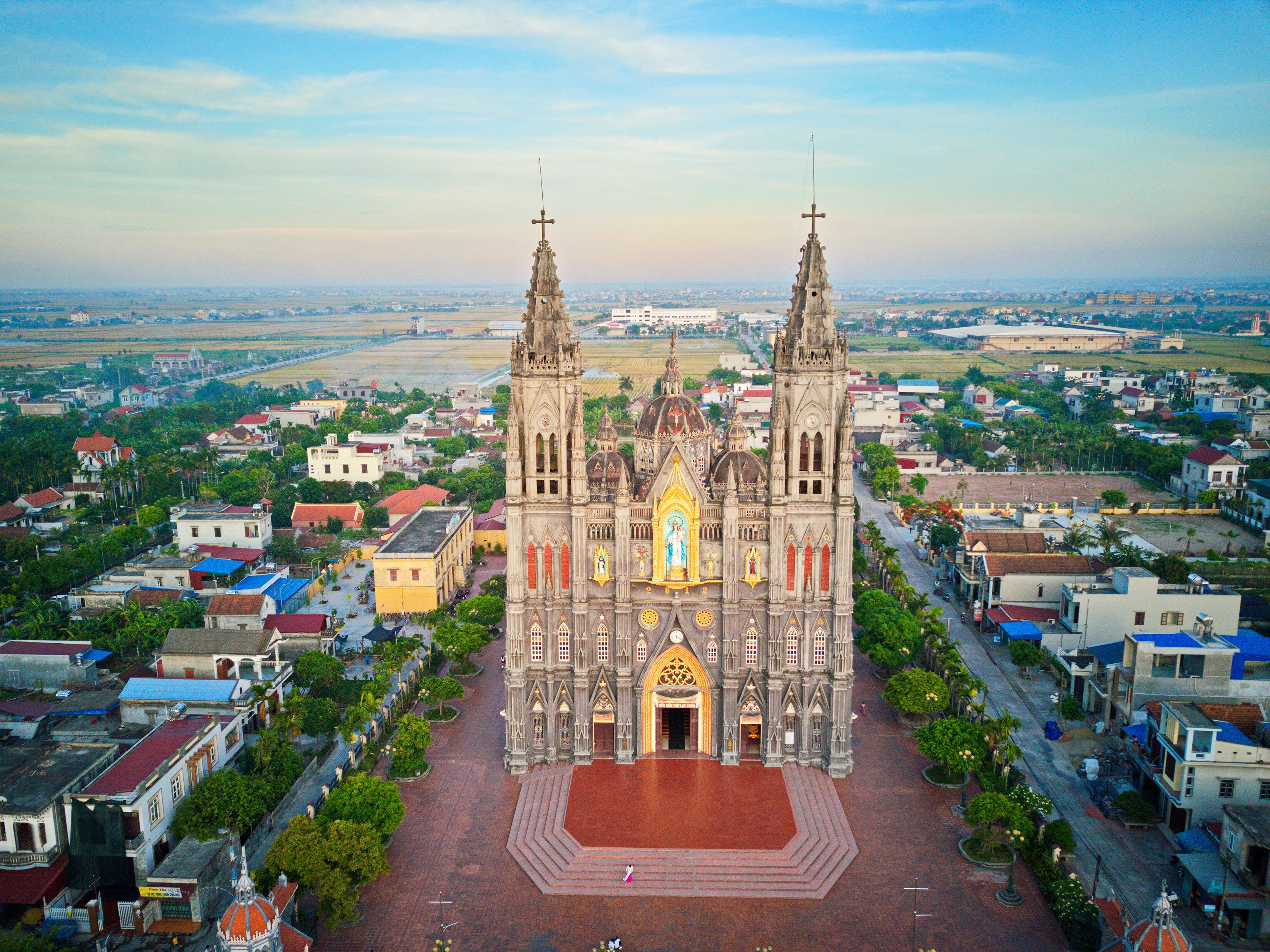A large cathedral with two towers and a central dome, surrounded by a town with various buildings and a distant view of fields.