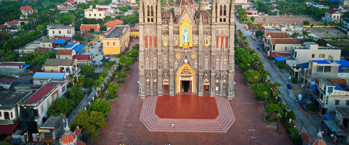 A large cathedral with two towers and a central dome, surrounded by a town with various buildings and a distant view of fields.