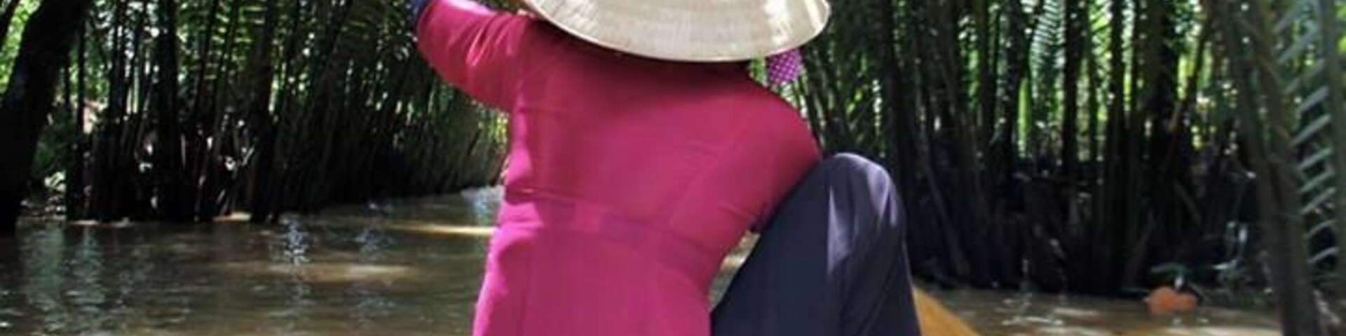 Una persona vestida de rosa y con sombrero de paja rema una barca por un bosque de manglares.