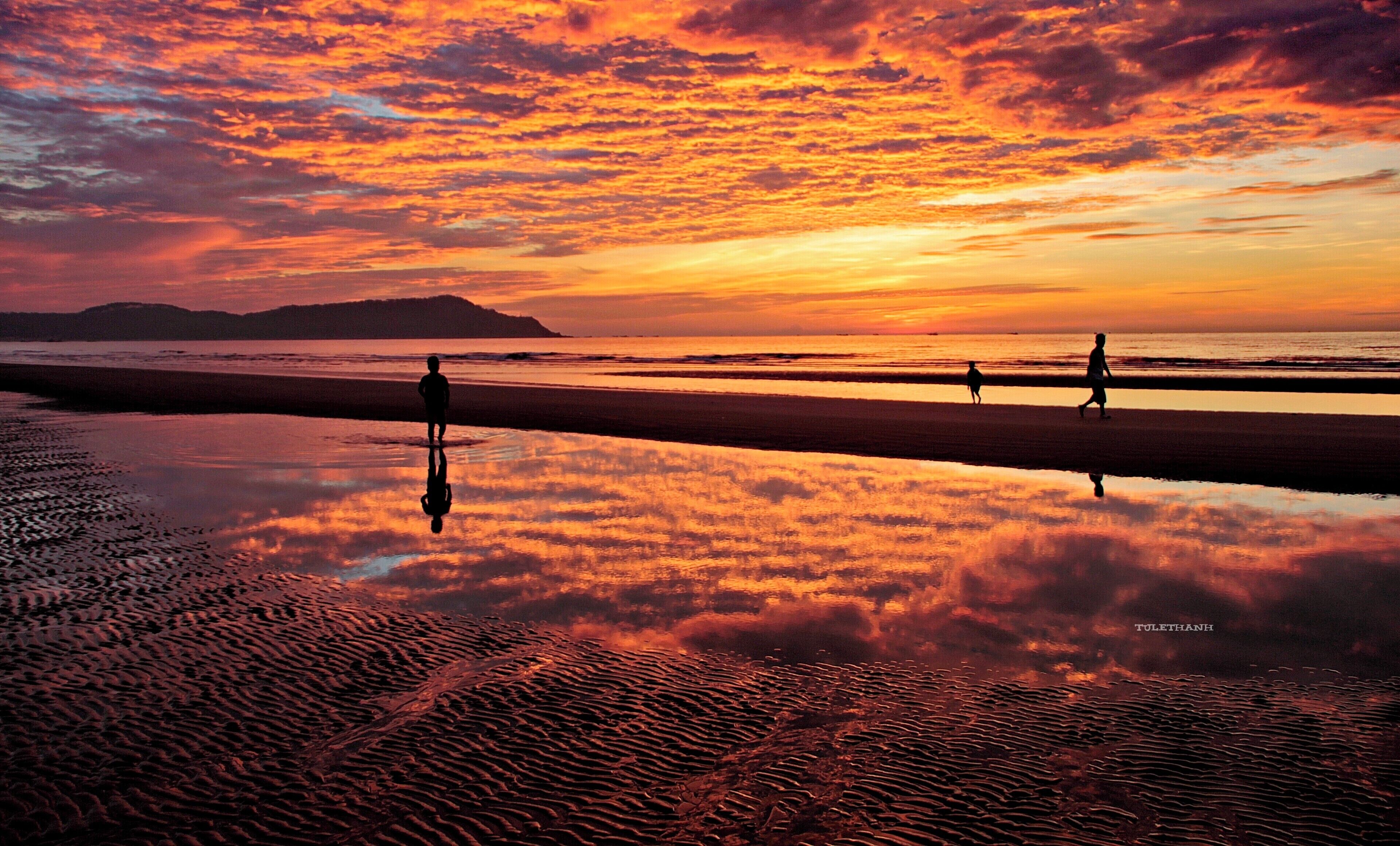 A beach at sunset with a vibrant sky, people walking, and a calm sea reflecting the colors.