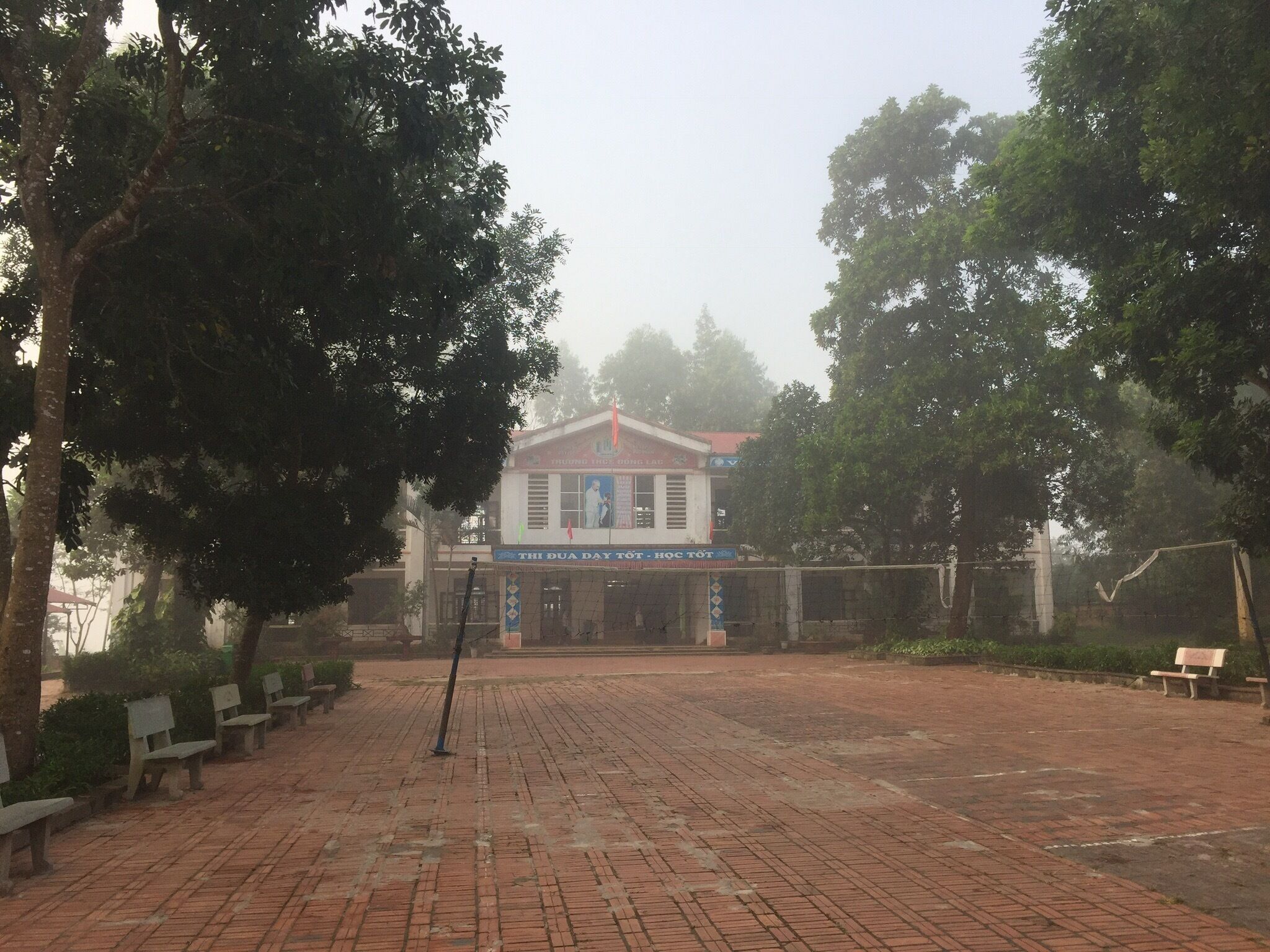 A brick-paved courtyard with benches, trees, and a building with a sign reading "THI DUA DAY TÔI HÒC TÔI".