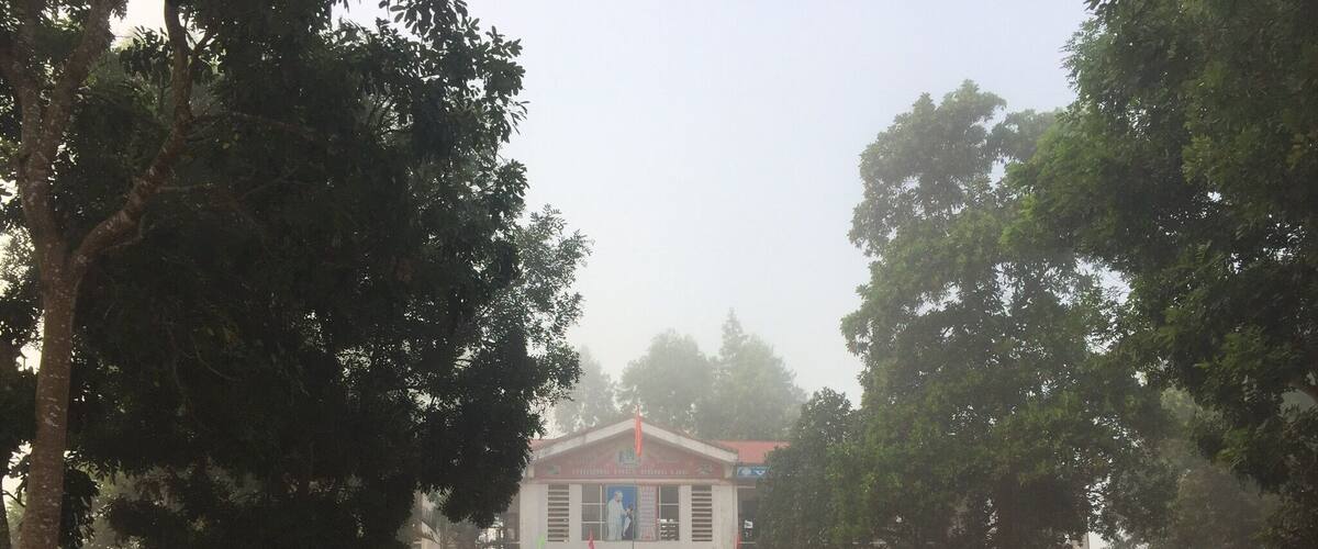 A brick-paved courtyard with benches, trees, and a building with a sign reading "THI DUA DAY TÔI HÒC TÔI".
