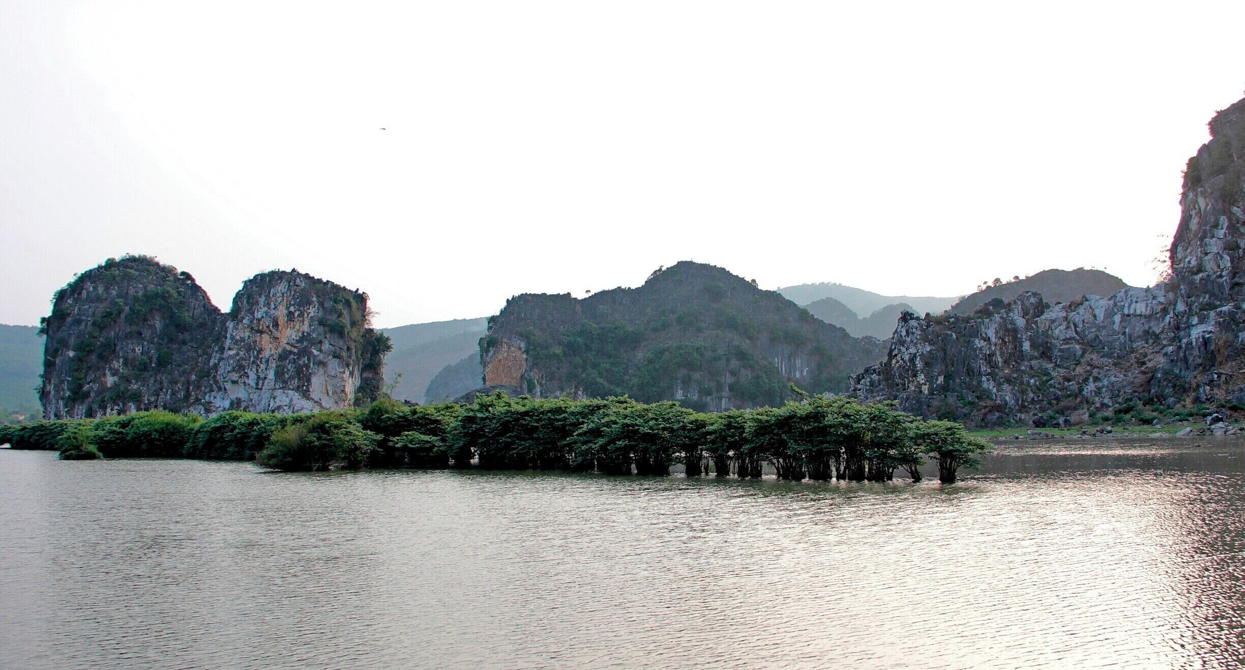 A calm river with rocky cliffs and a cluster of trees in the foreground.