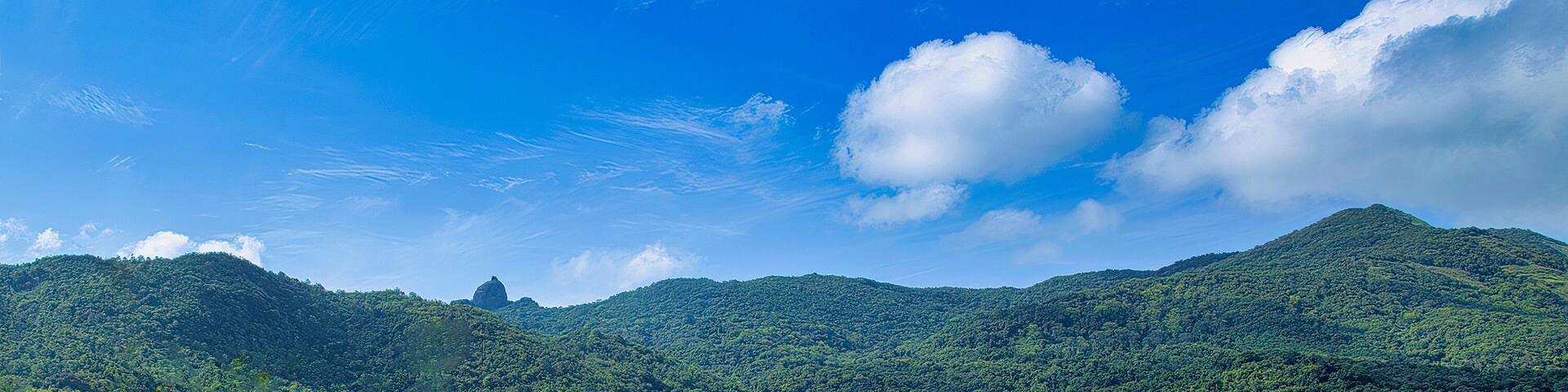 A mountainous landscape with dense green forests and a clear blue sky with scattered clouds.