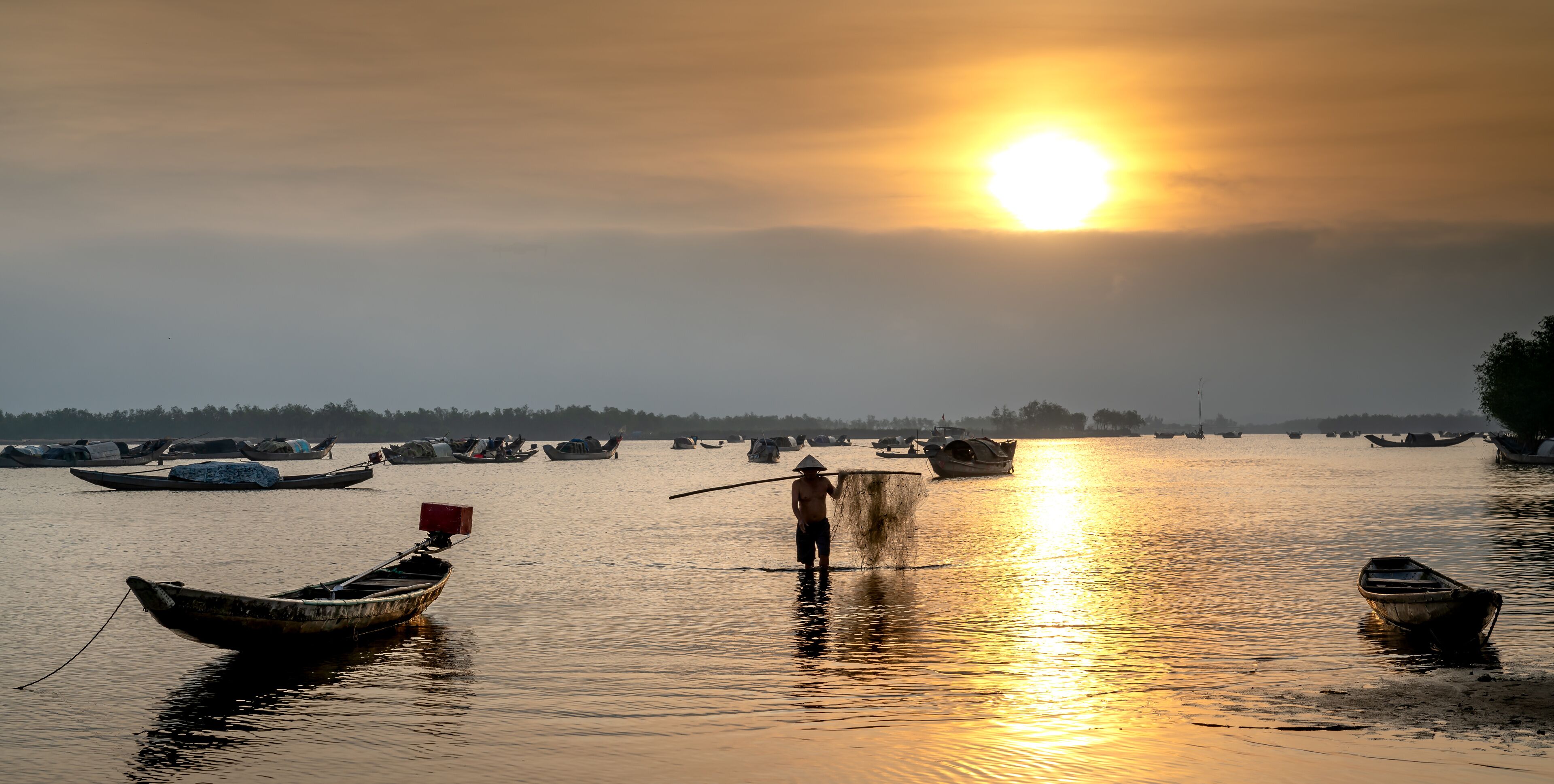 A calm river at sunset with several boats and a person standing in the water.