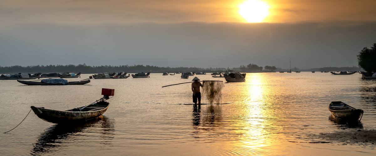 A calm river at sunset with several boats and a person standing in the water.