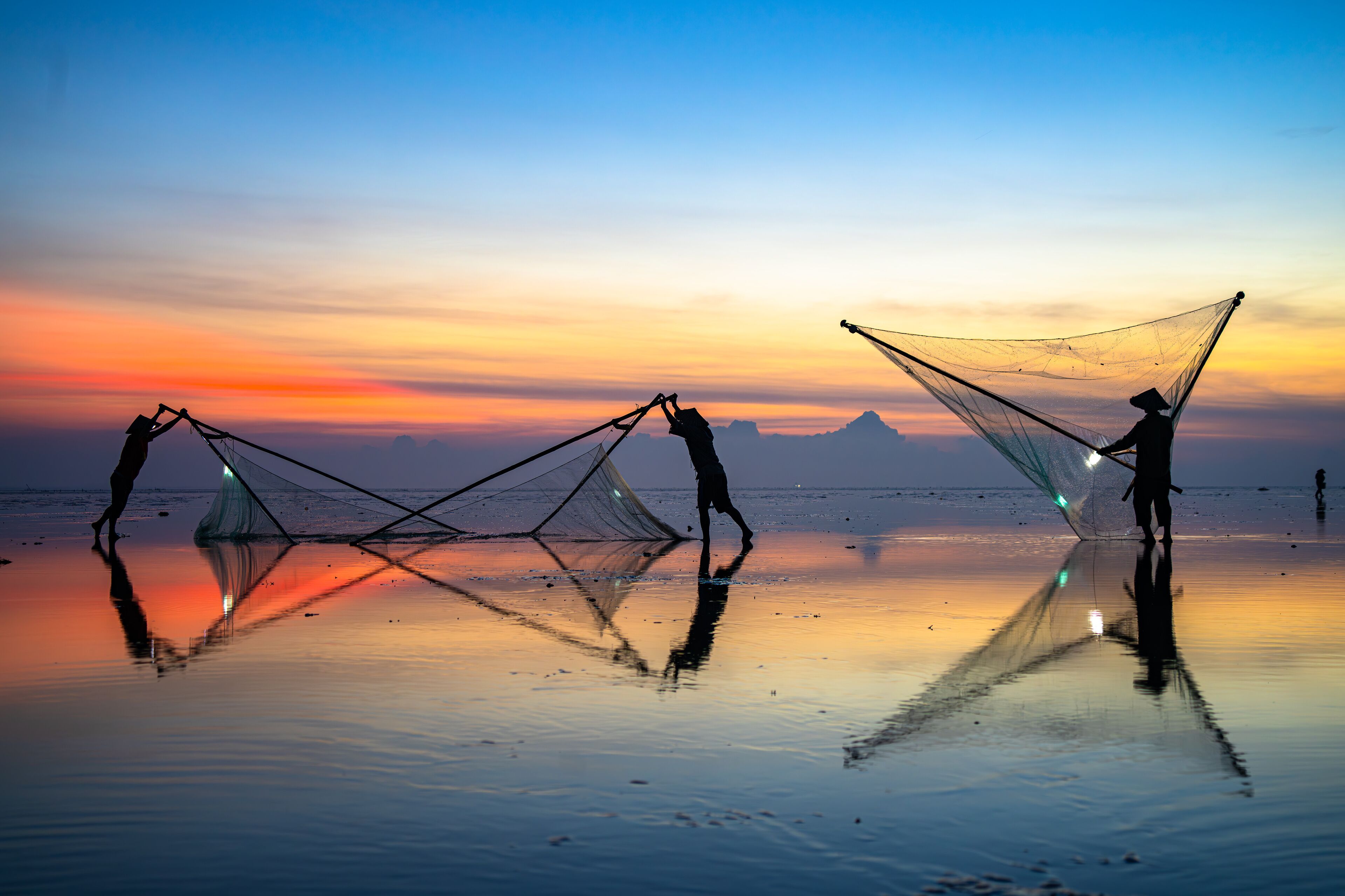 Two people casting fishing nets on a beach at sunset with mountains in the background.