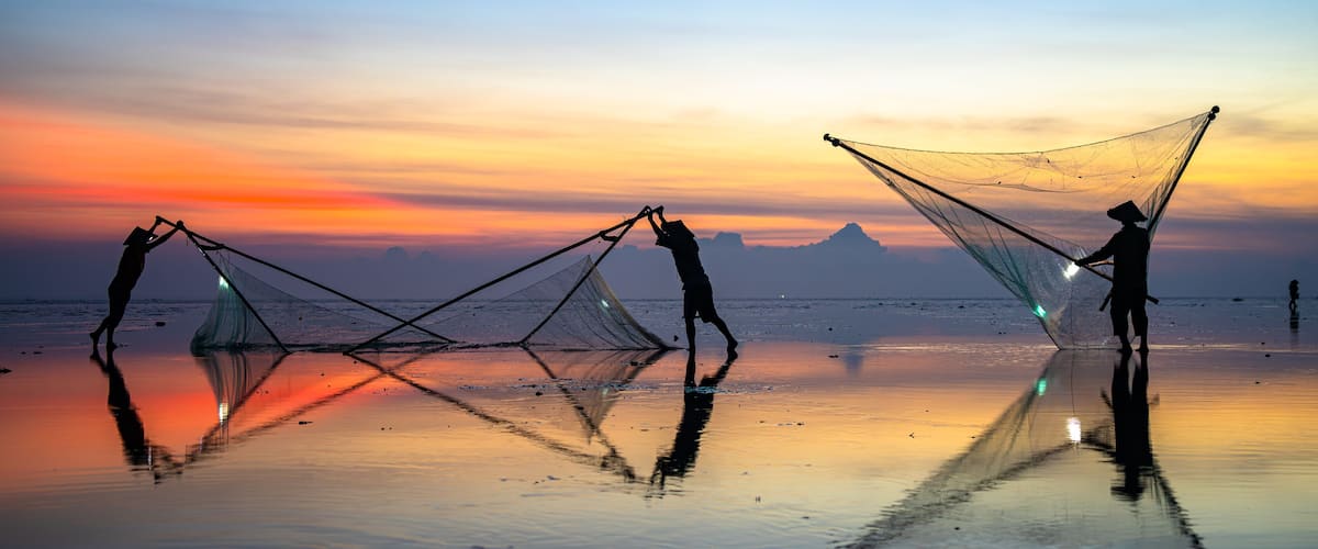 Two people casting fishing nets on a beach at sunset with mountains in the background.