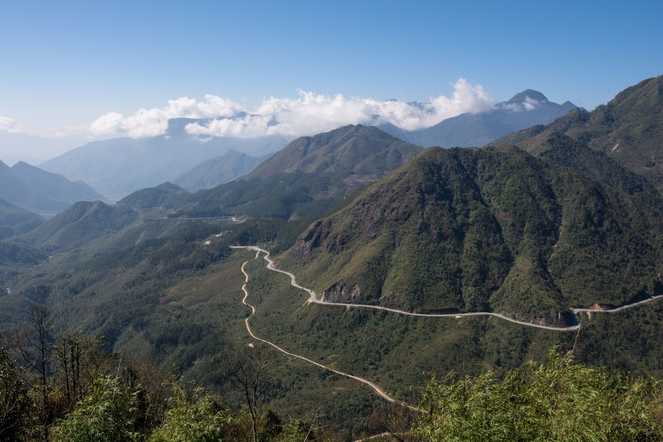 A winding mountain road through a lush, green valley with distant mountain peaks and clouds.