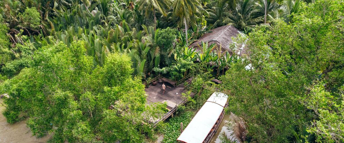 man standing on dock with boat on tropical coastline of Ben Tre along brown Mekong River with coconut tree jungle, aerial