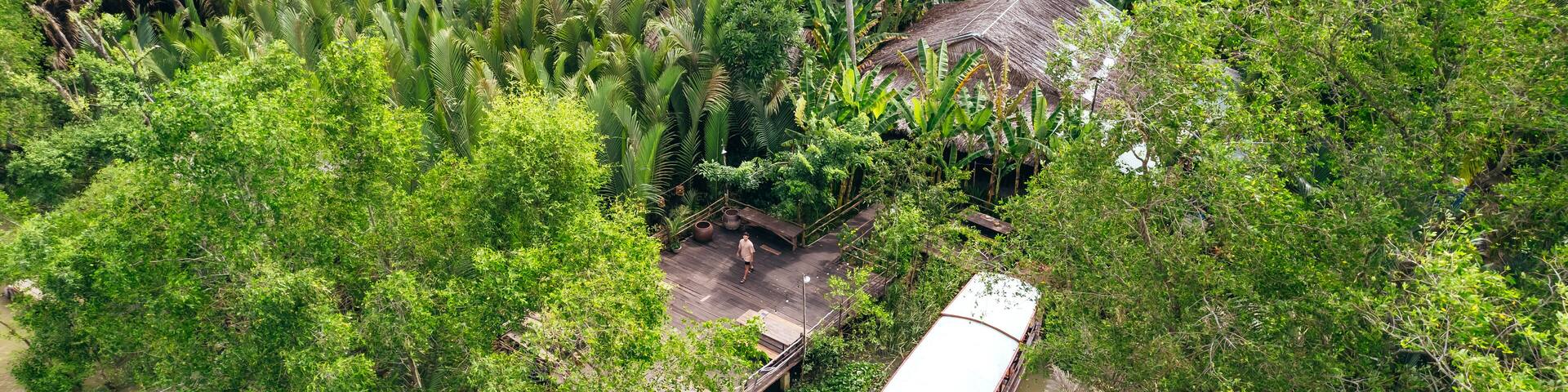 man standing on dock with boat on tropical coastline of Ben Tre along brown Mekong River with coconut tree jungle, aerial