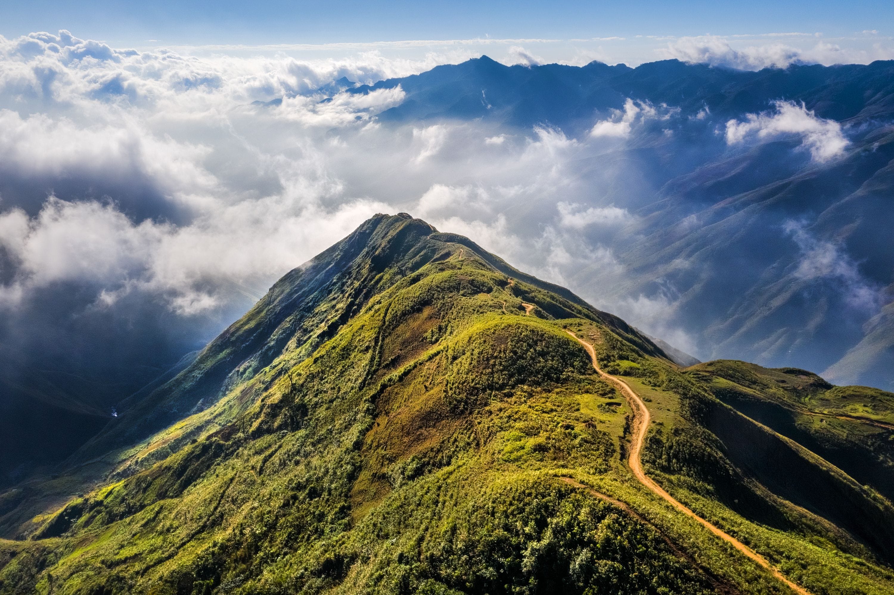 Un pico montañoso con un camino sinuoso que lleva a la cima, rodeado de nubes y otras montañas más bajas.