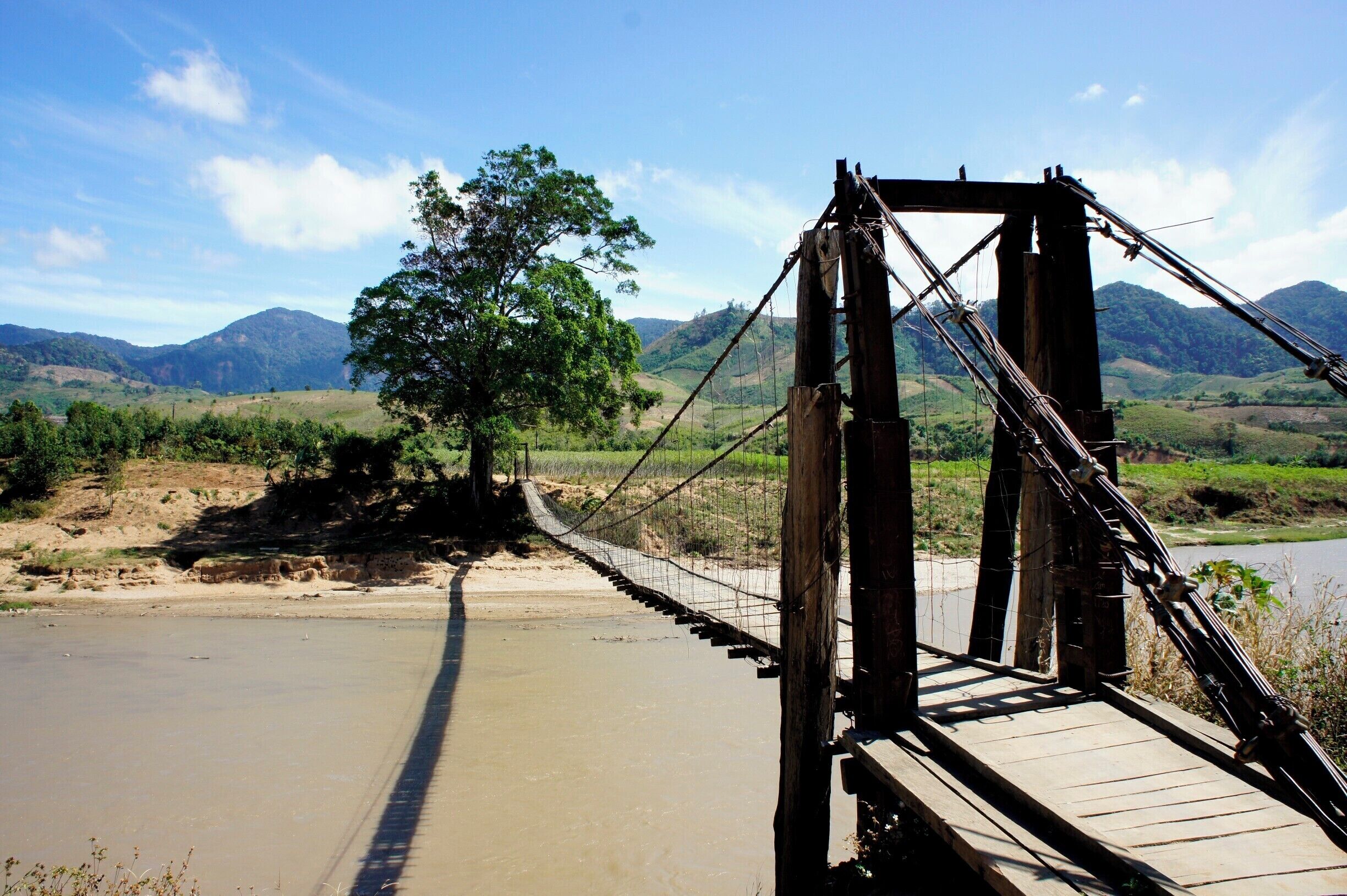 A wooden suspension bridge over a river with mountains in the background.